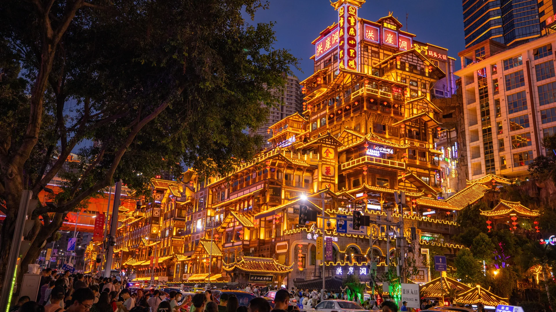 Hongya Cave, el complejo ribereño de estilo palafito en el centro de Chongqing, iluminado de dorado al atardecer con un árbol en primer plano. Es un lugar distinto del teatro.