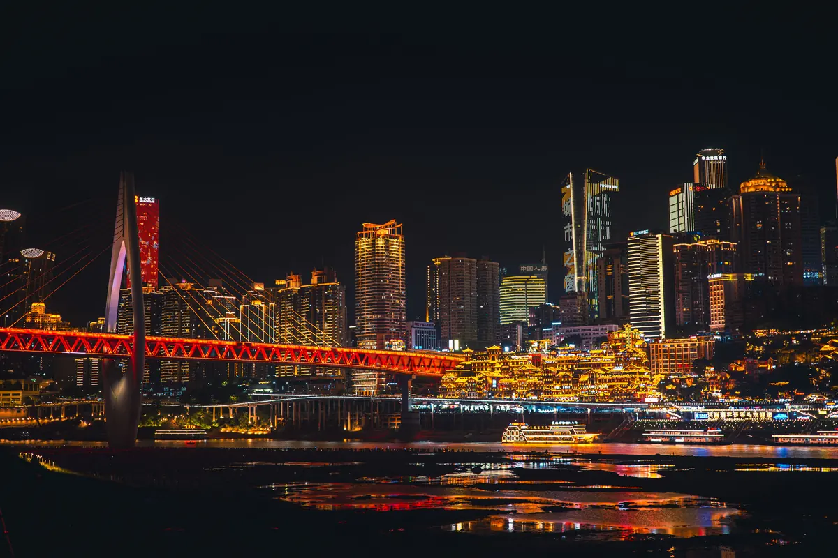 El horizonte de Chongqing al otro lado del río de noche, con un puente atirantado rojo y el complejo ribereño iluminado en oro.