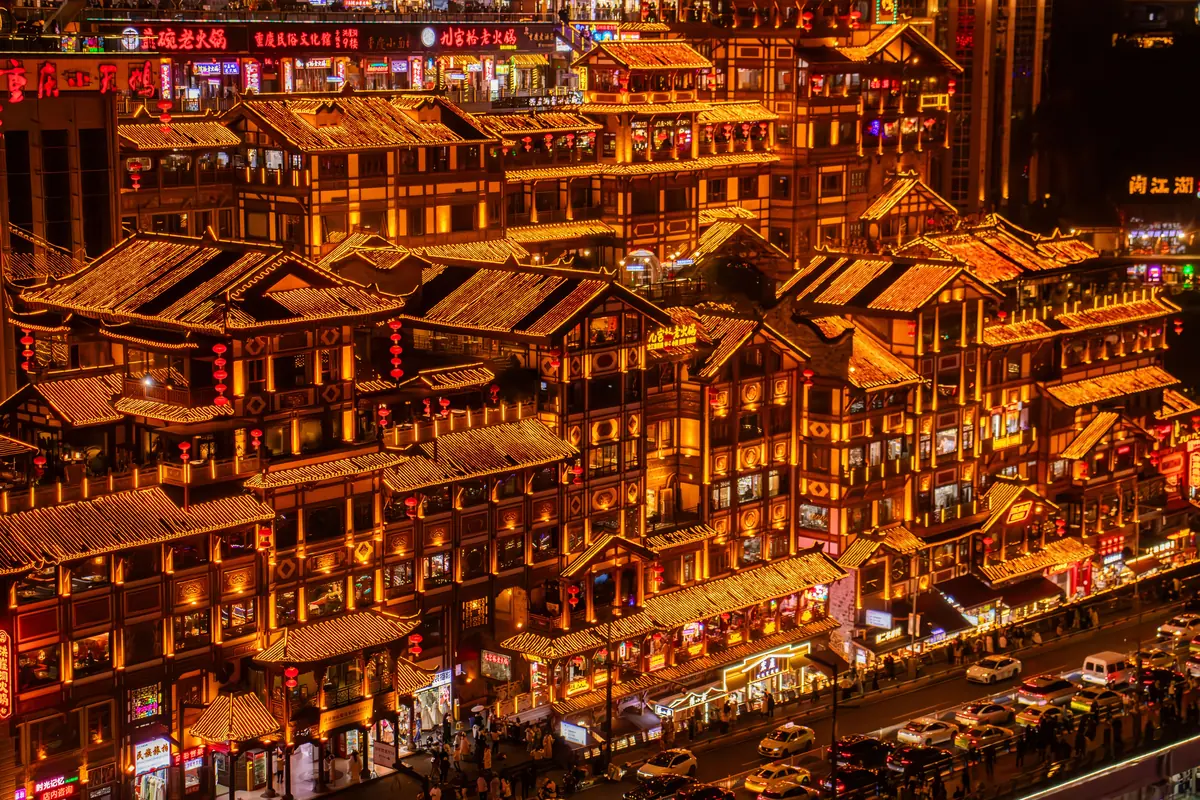 Niveles de aleros iluminados en oro y farolillos rojos en el paseo fluvial de estilo palafito de Chongqing de noche.