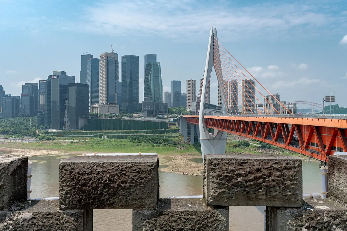 Un puente atirantado con tablero rojo que cruza el río en Chongqing, con torres detrás, visto entre almenas de piedra